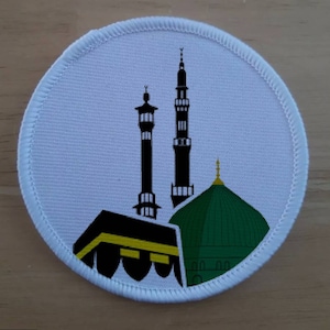 Puede incluir: Un parche redondo blanco con una silueta negra de la Kaaba, la mezquita de La Meca, Arabia Saudita. La Kaaba está rodeada por dos minaretes y una cúpula verde.