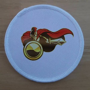 以下が含まれることがあります： 剣と盾を持った戦士の赤と金の刺繍デザインが施された白い丸いパッチ