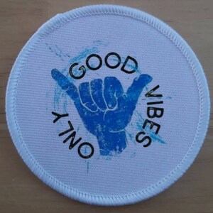 Puede incluir: Un parche redondo blanco con un gráfico de gesto de mano azul y las palabras "GOOD VIBES ONLY" en negro. El parche tiene un borde cosido y un diseño de salpicaduras azul claro.