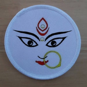 Puede incluir: Parche blanco bordado con una representación estilizada de la cara de una mujer. La cara tiene cejas negras, ojos negros grandes, un aro de nariz rojo y un diseño rojo y blanco en la frente.