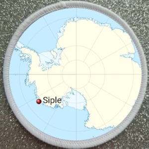 Può includere: Una toppa rotonda con una mappa dell'Antartide in azzurro e beige, con la parola "Siple" contrassegnata da un punto rosso. La toppa ha un bordo bianco, probabilmente ricamato o stampato.