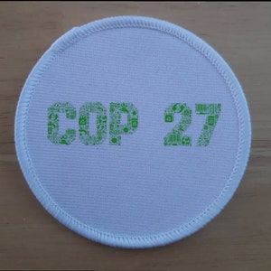 May include: White round fabric patch with green text that reads "COP 27". The text is made up of small green icons related to the environment.