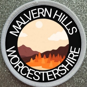 May include: A white round patch with black text and a black circle in the centre. The text reads "Malvern Hills Worcestershire". The circle contains a stylised image of a landscape with hills, trees, and a sun.