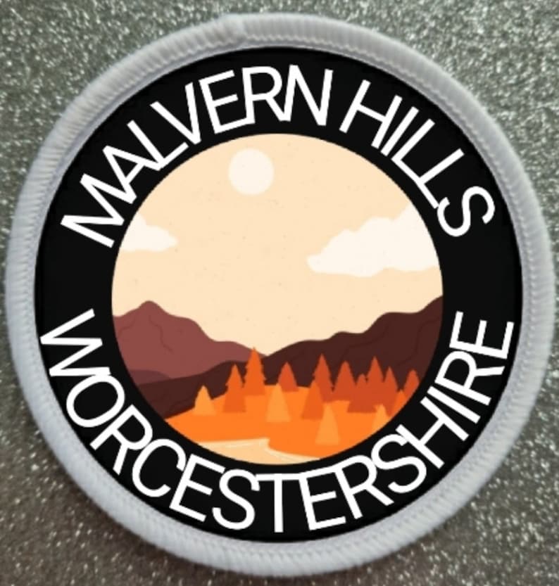 Peut inclure: Un &eacute;cusson rond blanc avec du texte noir et un cercle noir au centre. Le texte indique "Malvern Hills Worcestershire". Le cercle contient une image stylis&eacute;e d'un paysage avec des collines, des arbres et un soleil.