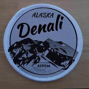 Puede incluir: Parche redondo blanco con una imagen en blanco y negro de la montaña Denali en Alaska. El texto "Alaska Denali 6190M" está impreso en el parche.