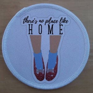 Könnte beinhalten: Runder weißer Stoff-Patch mit roten Glitzer-Schuhen und blauen Socken. Der Text "there's no place like HOME" ist in Schwarz gedruckt.