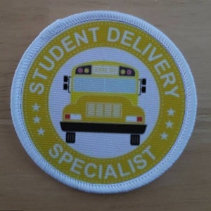 Może przedstawiać: Okrągła naszywka w kolorze żółtym i białym z tekstem "Student Delivery Specialist" w kółku wokół ilustracji żółtego autobusu szkolnego.