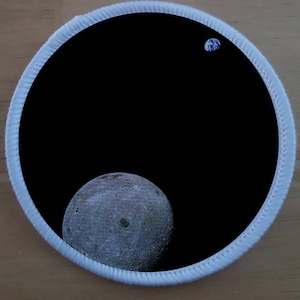 Könnte beinhalten: Ein schwarz-weißes Patch mit weißem Rand. Das Patch zeigt einen großen, grauen Mond mit Kratern und eine kleinere, blaue Erde in der oberen rechten Ecke.