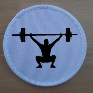 Puede incluir: Un parche redondo blanco con una silueta negra de una persona levantando una barra.