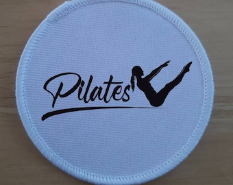 May include: White round fabric patch with black stitching around the edge. The patch features a black silhouette of a person doing a Pilates exercise and the word "Pilates" in a black cursive font.