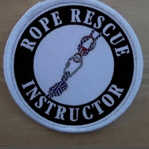 Könnte beinhalten: Runder Aufnäher mit dem Text "ROPE RESCUE INSTRUCTOR" in Weiß auf schwarzem Hintergrund. In der Mitte befindet sich eine detaillierte Abbildung eines Seilrettungssystems mit Karabiner und Knoten.