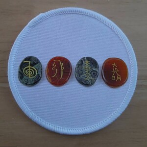 May include: Four oval-shaped stones with gold symbols and Japanese characters on a white background. The stones are green, red, gray, and red. The symbols are the Reiki symbol, the symbol for "joy", the symbol for "peace", and the symbol for "great light".