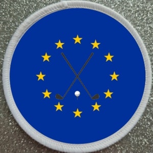 Op de afbeelding: Een ronde witte patch met een blauwe cirkel in het midden. De cirkel heeft een ontwerp van gekruiste golfclubs met een golfbal op de kruising. Er zijn 12 gele sterren in een cirkel rond de golfclubs.