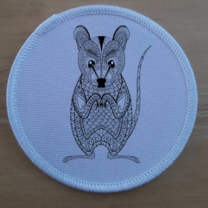 Op de afbeelding: Een ronde, witte patch met een gedetailleerde zwart-wit illustratie van een quokka. De quokka wordt afgebeeld in een staande pose met gevouwen handen. De patch heeft een witte gestikte rand.
