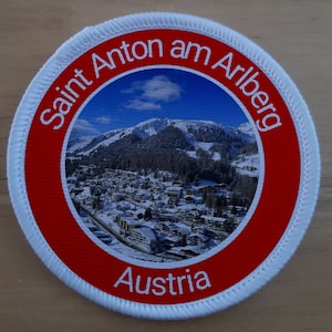 Pode incluir: Um remendo circular branco e vermelho com o texto "Saint Anton am Arlberg Áustria" e uma foto de uma vila de montanha com neve.