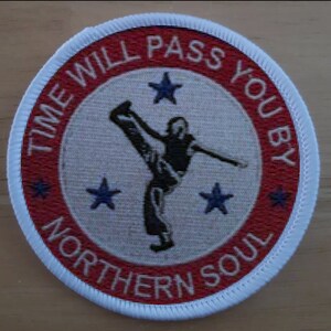 Op de afbeelding: Een ronde rode en witte patch met de tekst "Time Will Pass You By Northern Soul" rond de rand. In het midden van de patch is een zwarte silhouet van een persoon die een trap uitdeelt. Er zijn vijf blauwe sterren rond de persoon.