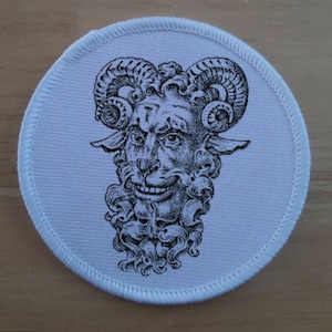 May include: A circular white patch featuring a black and white illustration of a horned face with a smiling expression. The face has a beard and curly hair, with a stitched border.
