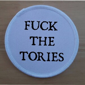 Peut inclure: Un écusson rond blanc avec le texte noir « FUCK THE TORIES ». L'écusson a une bordure blanche cousue et est probablement en tissu. Le texte est centré et en gras.