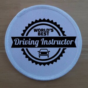 May include: White round fabric patch with black text and a car silhouette. The text reads "World's Best Driving Instructor".