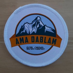 May include: White round patch with a blue and brown mountain scene. The text "AMA DABLAM" is in a brown banner across the bottom of the image. The text "6170m/20243ft" is below the banner.