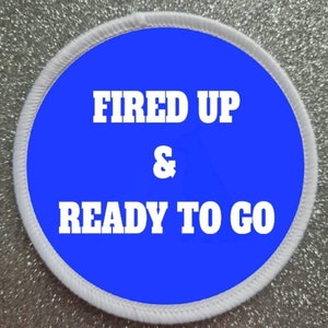 May include: A white round patch with a blue circle in the centre. The text "FIRED UP & READY TO GO" is written in white on the blue circle.