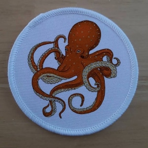 May include: A white round patch with a brown octopus with white spots on a white background.