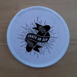 May include: White round patch with a black and white graphic of a skateboard with the text "Skate or Die" on a banner.