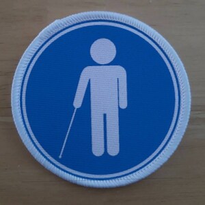 May include: A white circle with a blue border and a white stick figure holding a white cane in the centre. The circle is on a light brown wooden surface.
