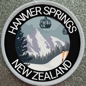 Puede incluir: Un parche redondo blanco con texto negro y un círculo negro en el centro. El texto dice "HAMMER SPRINGS NEW ZEALAND". El círculo contiene una escena de montaña nevada con telesillas y esquiadores.