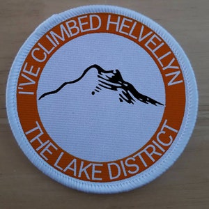 May include: White round patch with an orange circle in the center. The orange circle has the text "I've Climbed Helvellyn The Lake District" in white. A black silhouette of a mountain is inside the orange circle.