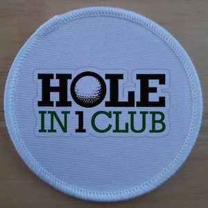 Könnte beinhalten: Runder weißer Patch mit einem schwarzen und grünen Design. Der Text "HOLE IN 1 CLUB" ist auf dem Patch mit einer Golfballgrafik in der Mitte des Wortes "HOLE" gedruckt.