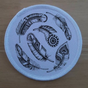 May include: White round fabric patch with black and white detailed feather designs. The feathers are arranged in a circular pattern.