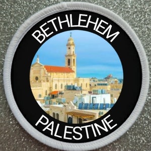 Può includere: Un cerotto circolare bianco con bordo nero e un cerchio nero al centro. Il cerchio nero contiene una foto di una chiesa a Betlemme, Palestina. Il testo "BETHLEHEM" è scritto in bianco sopra la foto e "PALESTINE" è scritto in bianco sotto la foto.