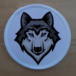 May include: A white circular patch featuring a black and grey wolf's head design. The wolf has blue eyes and a black nose.