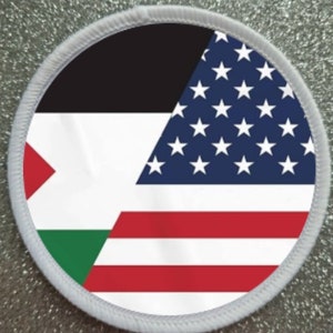 Pode incluir: Um remendo branco redondo com uma borda branca. O remendo apresenta as bandeiras da Palestina e dos Estados Unidos da América. A bandeira palestina está no lado esquerdo do remendo e a bandeira americana está no lado direito do remendo.