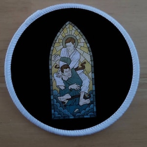 Puede incluir: Un parche redondo con borde blanco y fondo negro que presenta un diseño de vitral. La ventana representa dos figuras en una pose de lucha, una con un gi blanco y la otra con un gi verde, sobre un fondo azul y amarillo.