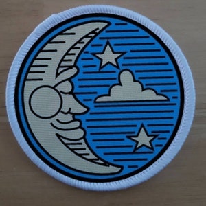 May include: A white, round patch with a blue background and a white crescent moon with a face. The moon is surrounded by white stars and a white cloud. The patch has a white border.