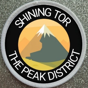 Op de afbeelding: Een ronde witte en zwarte patch met de tekst "Shining Tor The Peak District" in witte letters rond een zwarte cirkel met een gele en grijze bergillustratie erin.