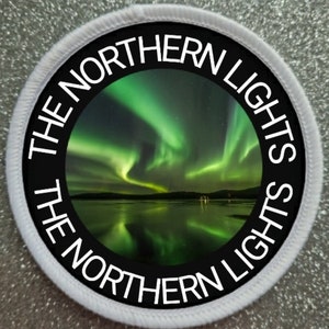 Könnte beinhalten: Ein runder bestickter Aufnäher mit einem Fotodruck des grünen Polarlichts über einer dunklen Landschaft und Wasser. Der schwarze Rand enthält den weißen Schriftzug THE NORTHERN LIGHTS, der zweimal um den Rand herum verläuft.