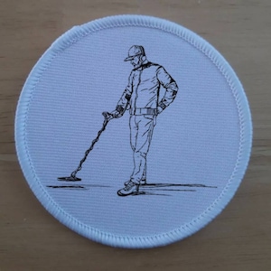 May include: A round, white patch with a black and white illustration of a person using a metal detector. The person is wearing a hat and jacket, holding the detector. The patch has a stitched border.