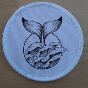 Può includere: Un cerotto rotondo bianco con un'illustrazione in bianco e nero di una coda di balena che emerge dalle onde.