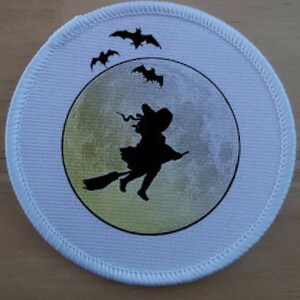 Pode incluir: Um remendo de tecido branco com uma silhueta preta de uma bruxa voando em uma vassoura na frente de uma lua amarela. Três morcegos pretos voam ao redor da lua.