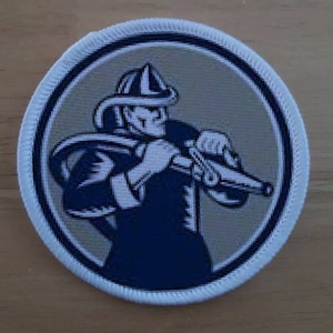 Könnte beinhalten: Ein weißer und blauer gestickter Aufnäher mit einem Feuerwehrmann, der einen Schlauch hält. Der Feuerwehrmann trägt einen Helm und hat einen entschlossenen Ausdruck im Gesicht.