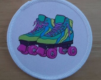 Puede incluir: Un parche redondo blanco con una ilustración colorida de un par de patines. Los patines son verdes, amarillos, azules y rosas. Las ruedas son rosas.