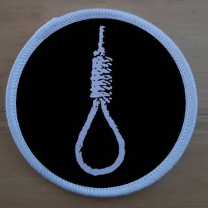 Può includere: Un cerotto circolare nero con bordo bianco, che rappresenta un cappio ricamato in bianco.