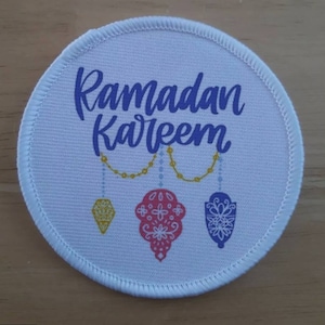 Puede incluir: Parche redondo de tela blanca con el texto "Ramadan Kareem" en letras cursivas azules. Debajo del texto se representan tres adornos colgantes: un diamante amarillo, un adorno estampado en rosa y blanco y un adorno estampado en azul y blanco.