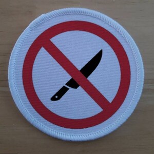 Peut inclure: Écusson rond blanc avec un symbole rouge d'interdiction sur un couteau noir. Le patch a une bordure blanche cousue. Le couteau est noir avec un point blanc sur le manche, indiquant une interdiction de couteaux.
