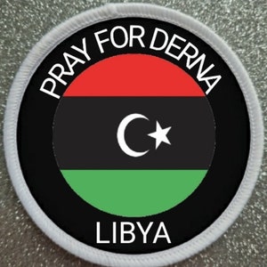 May include: A white circular patch with black and white text that reads "Pray for Derna Libya". The centre of the patch features the Libyan flag, which is a horizontal triband of red, black, and green with a white crescent moon and star.