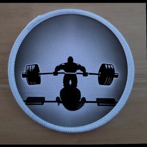 May include: A white circular patch with a black silhouette of a person lifting a barbell. The person is in a squat position with the barbell resting on their shoulders. The background is a gradient of light gray to dark gray.
