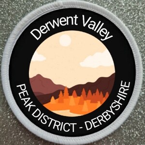 May include: Round patch with a landscape design. The text reads "Derwent Valley PEAK DISTRICT - DERBYSHIRE". The design features a sun, mountains, trees, and a river, in orange, brown, and beige, set against a black background.